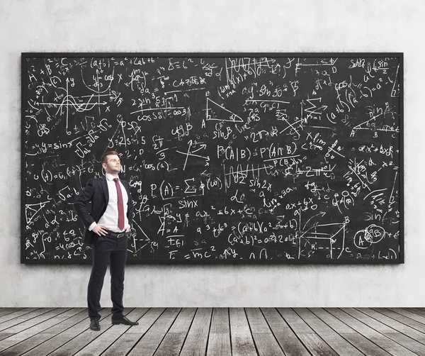Side view of a confident young man who stands in front of black chalkboard which is full of math formulas. A concept of Analyst at investment bank. — Stockfoto