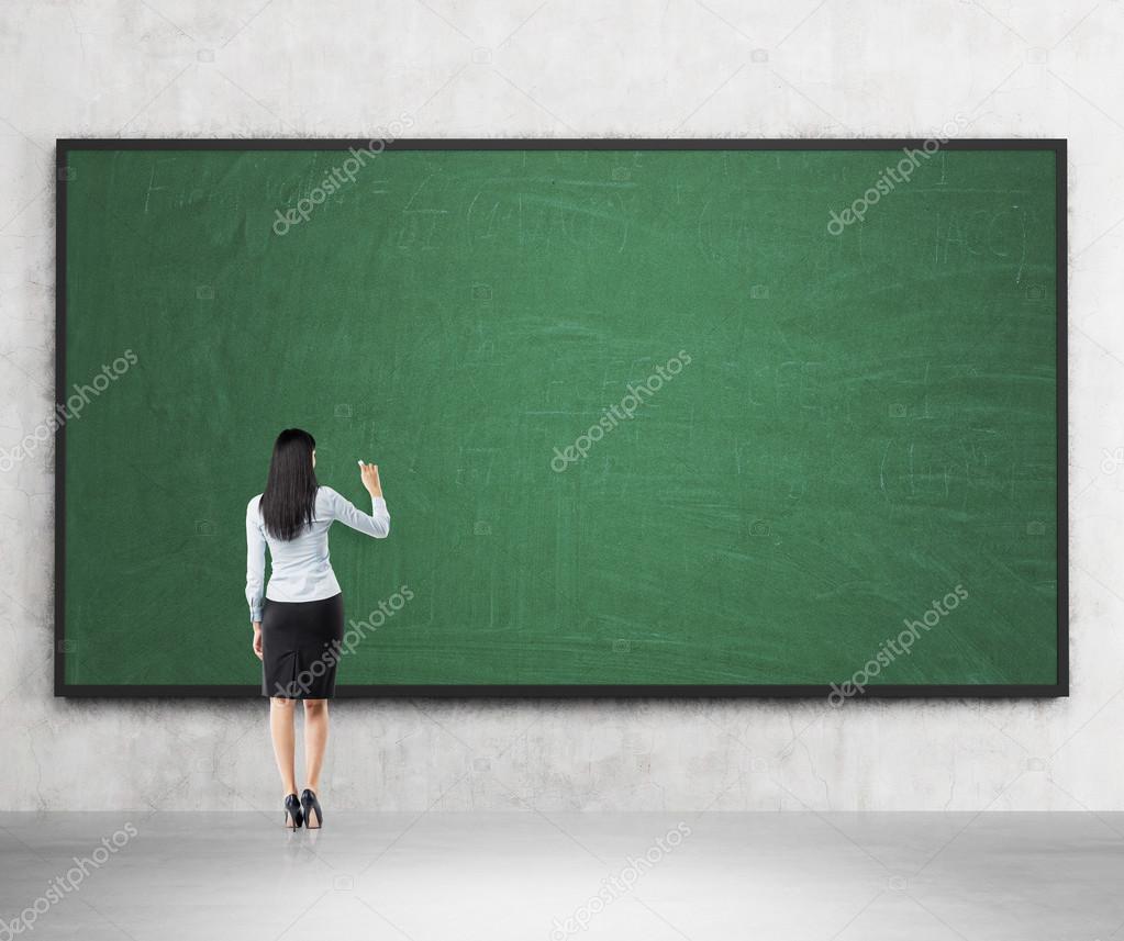 Full length of the rear view of the brunette girl who is going to write something on the green chalkboard. A concept of the beginning of new academic year. A class room.