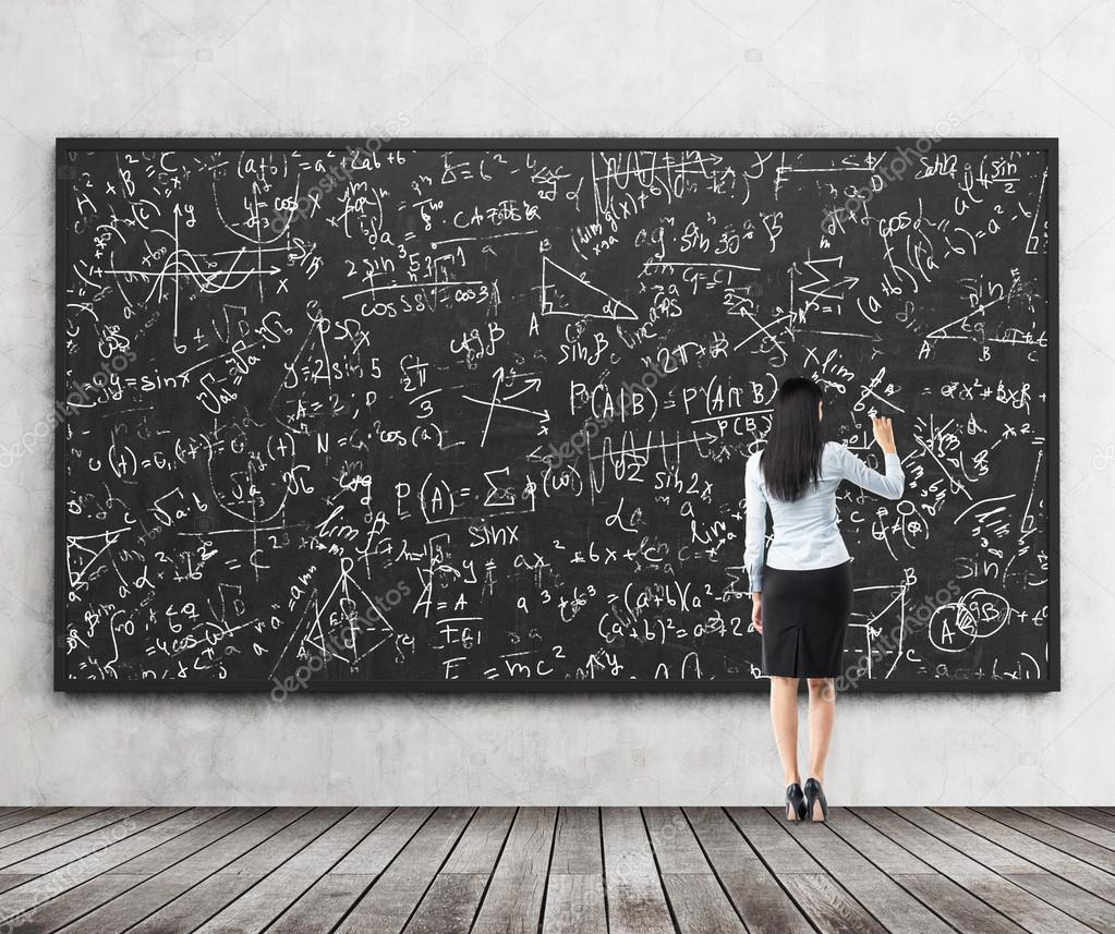 Full length of the rear view of the brunette girl who is writing down math formulas on the black chalkboard. A concept of university education.