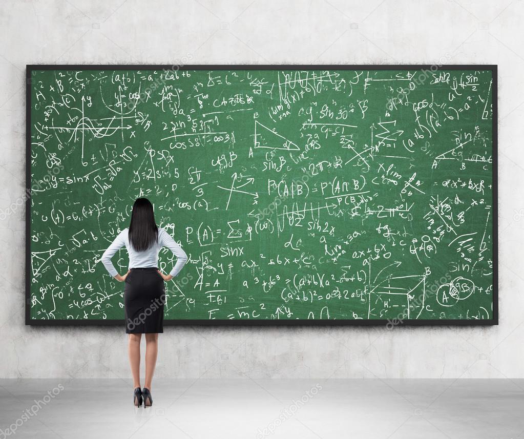 Full length of the rear view of the brunette lady who is looking at the math formulas which are drawn on the green chalkboard. A concept of university education.