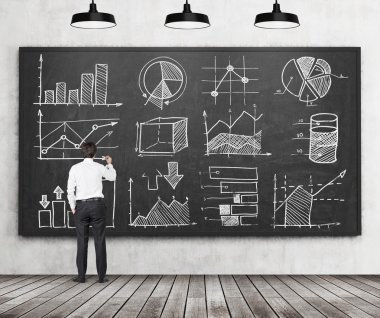 Young businessman or student of finance or management programme is drawing some charts or graphs on the black chalkboard. Rear view of the model. Wooden floor and three ceiling lights in the room.