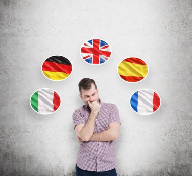 Young man in casual shirt holds his chin and thinks about which language to study. Italian, German, United Kingdom, Spanish and French flags in the bubbles are flying above the person.