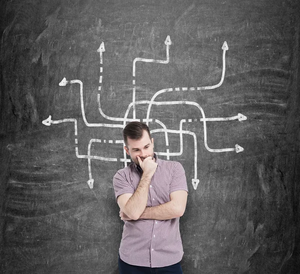 Young man in casual shirt holds his chin and thinks about the best solution of the problem. A sketch of the arrows in different directions are drawn on the black chalkboard. — Stockfoto