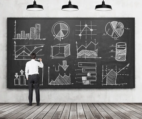 Young businessman or student of finance or management programme is drawing some charts or graphs on the black chalkboard. Rear view of the model. Wooden floor and three ceiling lights in the room. — 图库照片