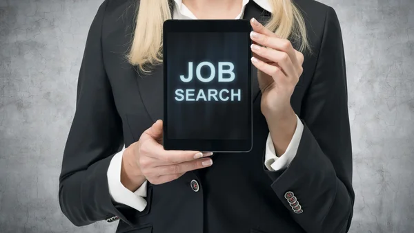 Blonde woman in formal suit presents a tablet with the words 'Job Search' on the screen. A concept of recruitment process. Internship and graduate programmes. Concrete background. — стокове фото