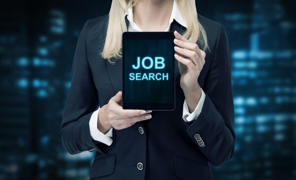 Blonde woman in formal suit presents a tablet with the words 'Job Search' on the screen. A concept of recruitment process. Lights of the evening business city in blur on the background.