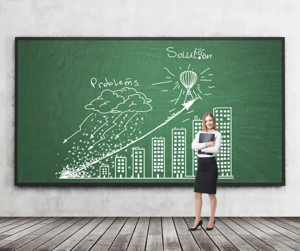 A brunette young woman in formal clothes holds a black folder and presents ' Problems and solution ' strategy with the graph on the green chalkboard. Wooden floor and concrete walls in the room.