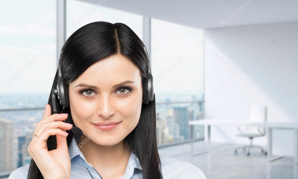 Front view of the smiling brunette support phone operator with headset. Panoramic office workplace on the background. New York city view.