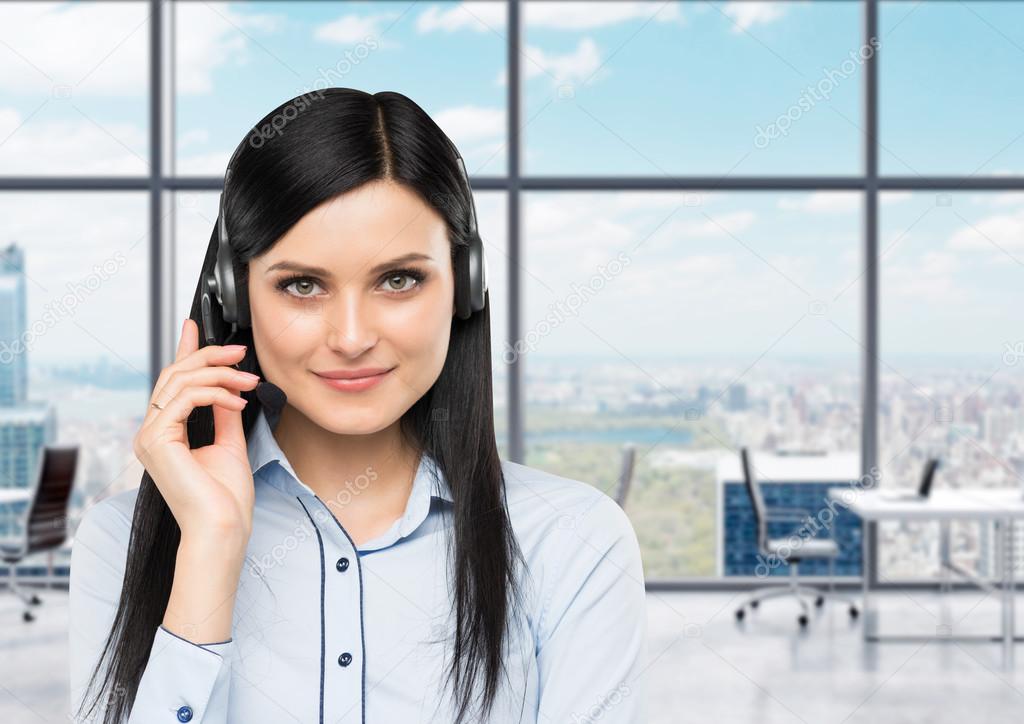 Front view of the smiling brunette support phone operator with headset. Office panoramic office on the background in blur. New York city panoramic view.