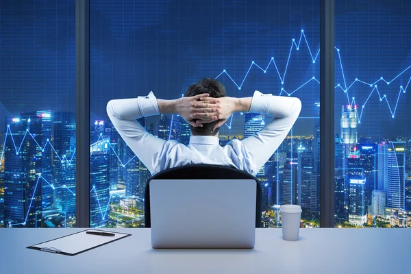 Задний вид сидящего бизнесмена, который смотрит на город из современного панорамного офиса. Нью-Йорк вечерний вид. Скрещенные руки на голове. Финансовые диаграммы нарисованы поверх панорамных окон . — стоковое фото