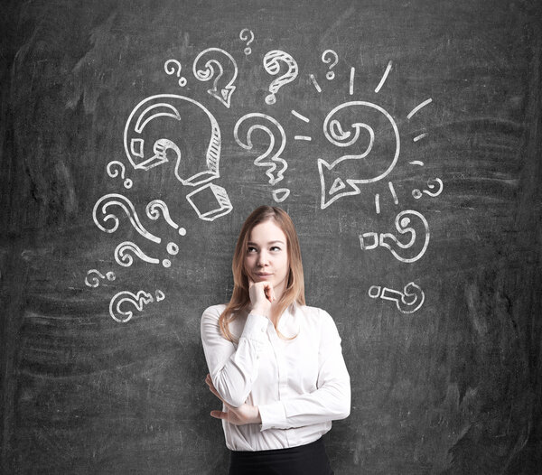Beautiful young woman in formal clothes holds her chin and thinks about unsolved problems. Question marks are drawn around the head. Black chalk board on the background.