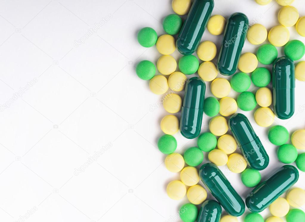 A top view of a heap of yellow and green medicine pills and capsules on white surface.