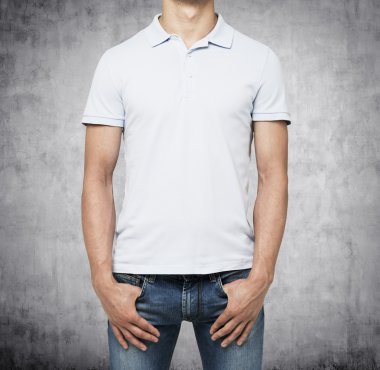 A man in a white polo shirt and denims holds his hands in pockets. Concrete background.