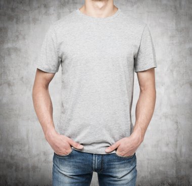 A man in a grey t-shirt and denims holds his hands in pockets. Concrete background.