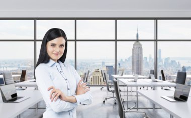 Smiling brunette business lady with cross hands is standing in a modern panoramic office in New York City. Manhattan panoramic view. Workplaces are on the office open space behind the model. clipart