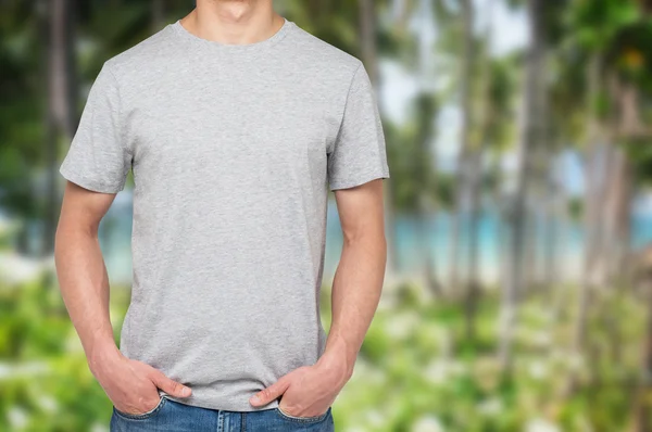 A man in a grey t-shirt and denims holds his hands in pockets. Tropical landscape and sea view in blur on the background. — Stok fotoğraf