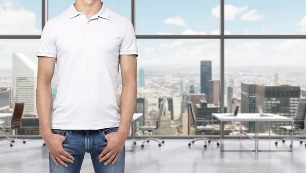 A man in a white polo shirt and denims holds his hands in pockets. A modern panoramic office space in New York City. Manhattan panoramic view. — Stock Photo, Image