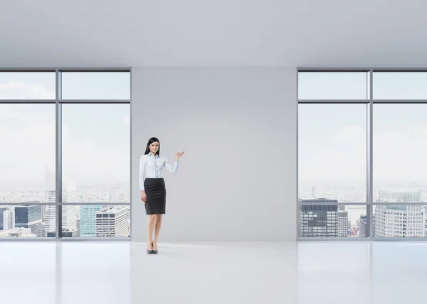 Ragazza bruna a figura intera in abiti formali sta indicando qualcosa per mano sulla parete bianca. Un moderno ufficio panoramico a Manhattan, New York . — Foto Stock