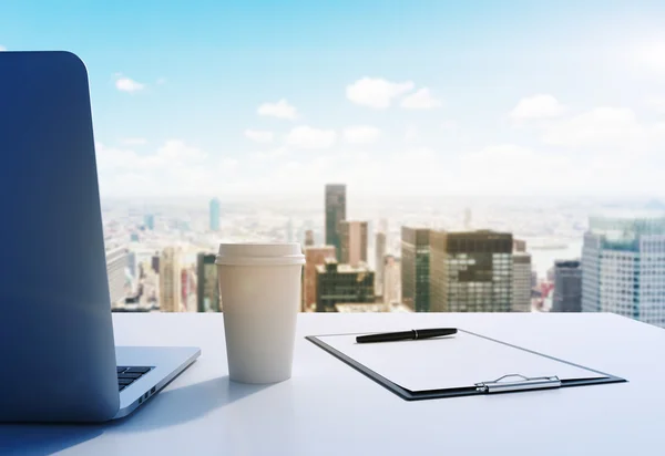 Un lugar de trabajo en una moderna oficina panorámica en Manhattan, Nueva York. Una computadora portátil, un bloc de notas y una taza de café están en la mesa blanca. Representación 3D. Imagen tonificada . — Foto de Stock