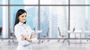 A side view of a beautiful smiling lady with crossed hands. The brunette woman is dressed in formal clothes. Modern workplaces in a fancy panoramic office in Singapore.
