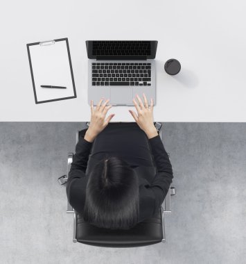 A top view of a brunette at the working desk with a laptop, a writing pad and a cup of coffee. Concrete floor.
