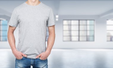 Front view of a man in a light grey t-shirt and denims. Modern loft style open space on the background.