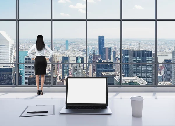 Una donna bruna vestita in abiti formali si trova in un moderno ufficio panoramico e guarda New York. Un computer portatile con schermo bianco, un blocco di scrittura e una tazza di caffè sono sul tavolo bianco . — Foto Stock