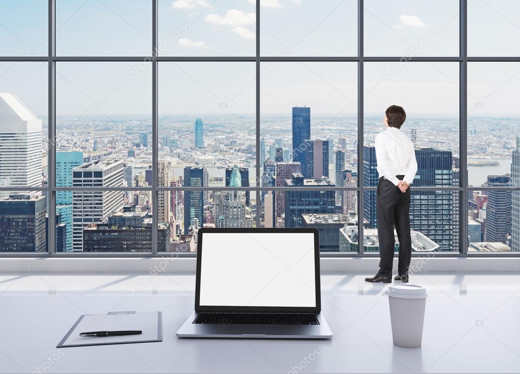 A dressed person in formal clothes is standing in modern panoramic office and looking at New York. A laptop with white screen, a writing pad and a cup of coffee are on the white table.