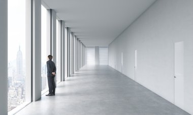 Young professional in formal suit is looking out the window. Empty modern bright clean interior of an open space office. Huge panoramic windows with New York view.