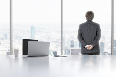 A young professional is looking out the window in the modern panoramic office in Manhattan, New York City. A laptop, notepad and a coffee cup are on the white table.