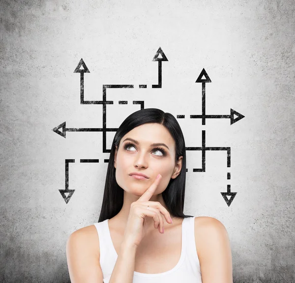 A beautiful brunette woman who is pondering about possible solutions of the complicated problem. Many arrows with different directions are drawn around her head. Concrete wall on background. — Stock fotografie