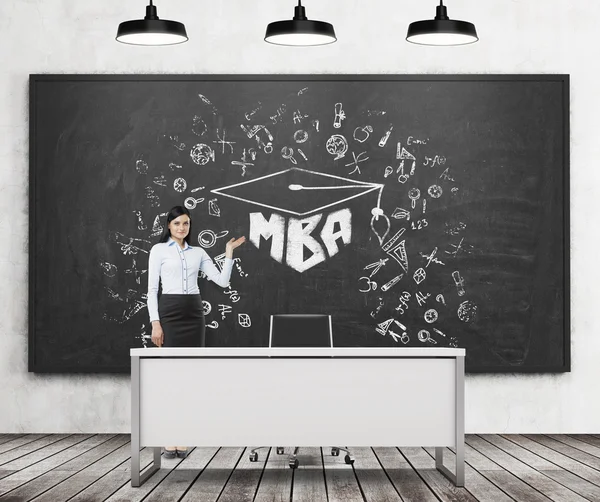 A brunette girl presents MBA sketch on the black chalk board in a modern classroom. Three black ceiling lights, wooden floor and concrete wall. A concept of business education. — Stock Fotó