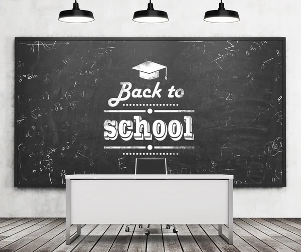 Teacher's desk in a modern university or school. A huge black chalkboard on the wall with written down phrase - Back to school. Three black ceiling lights and wooden floor. 3D rendering. — Stock Fotó