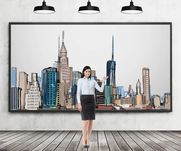 A brunette lady is pointing out the picture of New York City on the wall. Wooden floor, concrete wall and three black ceiling lights. — стокове фото