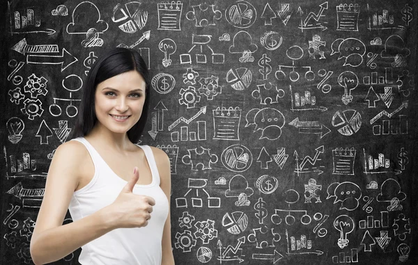 Brunette smiling woman with thumb up gesture. Business icons are drawn on the black chalk board on the background. — Stock Photo, Image