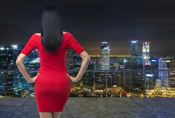 Vista trasera de la hermosa morena en vestido rojo posando en la terraza de la azotea moderna con panorámica de la noche Nueva York paisaje urbano . — Foto de Stock