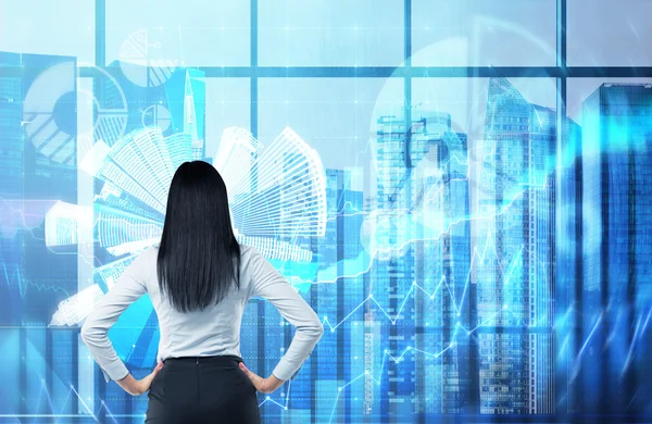 Rear view of the brunette woman who is looking out the window in the panoramic office in New York. Financial charts are over the whole panoramic windows. — Stock Fotó