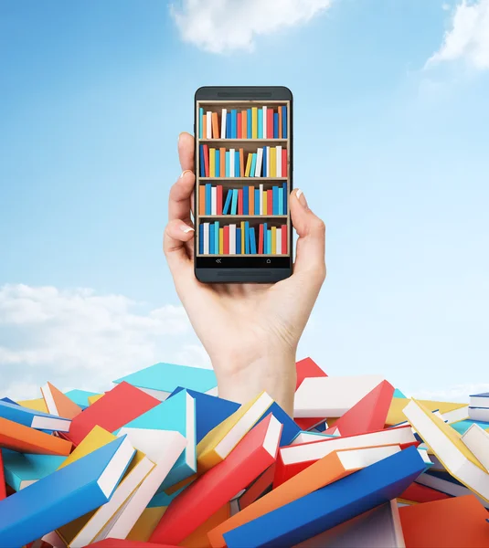 Una mano sostiene un teléfono inteligente con un estante de libros en la pantalla. Un montón de libros coloridos. Un concepto de educación y tecnología.Nublado cielo azul en el fondo . — Foto de Stock