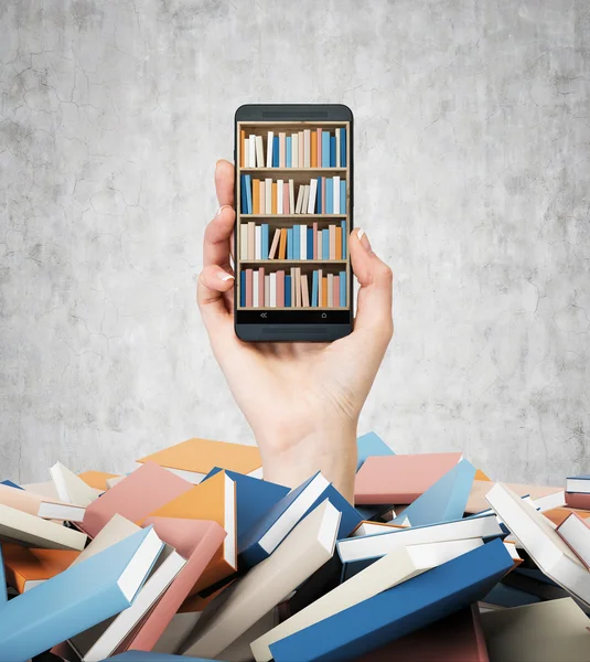 A hand holds a smartphone with a book shelf on the screen. A heap of colourful books. A concept of education and technology. Concrete wall background. — 스톡 사진