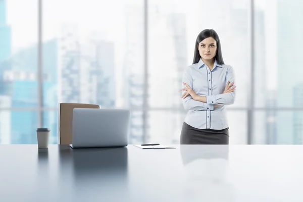 Eine junge brünette Dame mit verschränkten Händen im modernen Panorama-Büro in Singapore. Laptop, Notizblock und Kaffeetasse liegen auf dem weißen Tisch. — Stockfoto