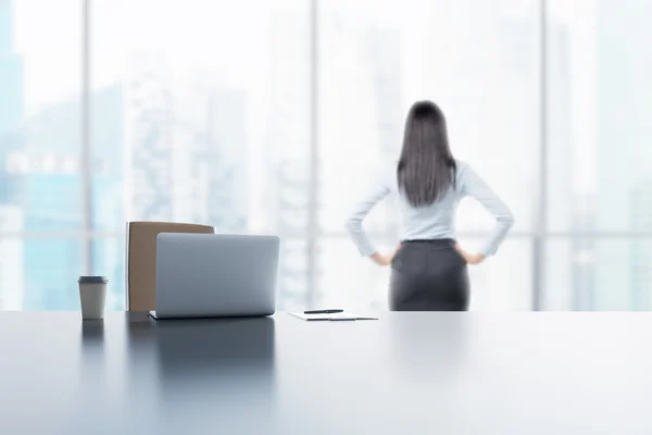 Una giovane bruna guarda fuori dalla finestra nel moderno ufficio panoramico di Singapore. Un computer portatile, blocco note e una tazza di caffè sono sul tavolo bianco . — Foto Stock