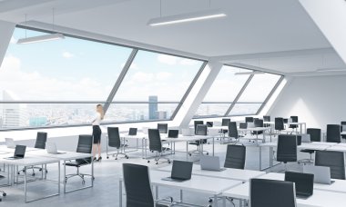 A young woman is looking out the window in the modern bright open space loft office. White tables equipped by modern laptops and black chairs. New York panoramic view. clipart