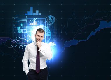 Handsome businessman is thinking about business development. Blue holograms of business charts are flying behind the person. Dark background.