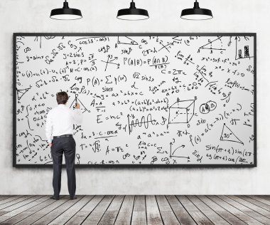 Rear view of young professional who is writing down math formulas on the whiteboard. A concept of analytical calculations. Wooden floor, concrete wall and three black ceiling lights are in the room.