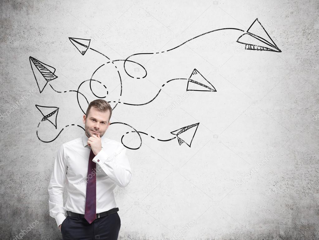 Handsome businessman is pondering about possible solutions of the complicated problem. Many arrows with different directions are drawn around his head on the concrete wall.