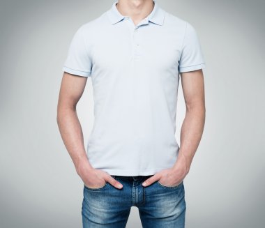 A man in a light blue polo shirt and denims holds his hands in pockets. Grey background.