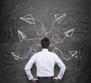 Rear view of a man in formal clothes who is pondering about possible solutions of the complicated problem. Many arrows with different directions are drawn around his head on the black chalkboard.