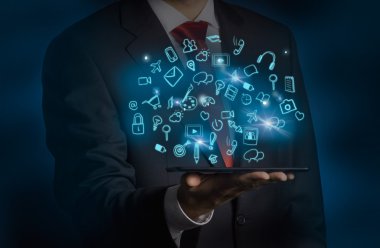 A man in formal suit is holding a tablet with flying out hologram social media icons. Dark background.