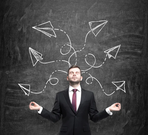 Meditative handsome businessman is pondering about possible solutions of the complicated problem. Many arrows with different directions are drawn around his head. Black chalkboard as a background. — Stock fotografie