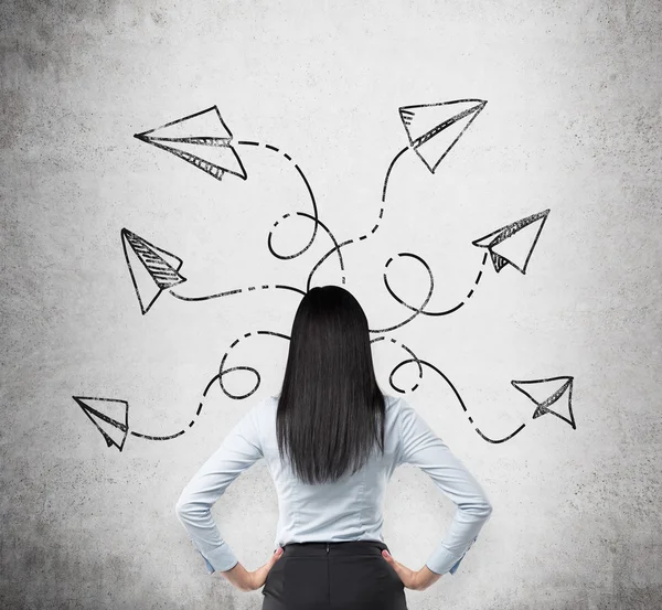 Rear view of brunette in formal clothes who is pondering about possible solutions of the complicated problem. Many arrows with different directions are drawn around her head on the concrete wall. — Stock Photo, Image